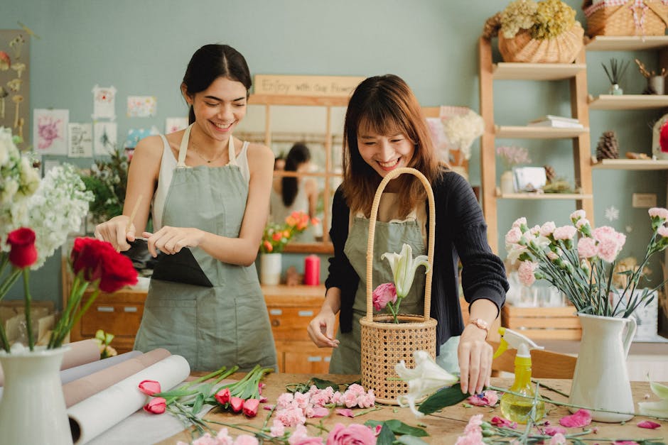 A florist and a customer inside a flower shop, with the florist wearing a light green apron over a white top and the customer in a dark sweater, both focused on a laptop placed on a rustic wooden tabl