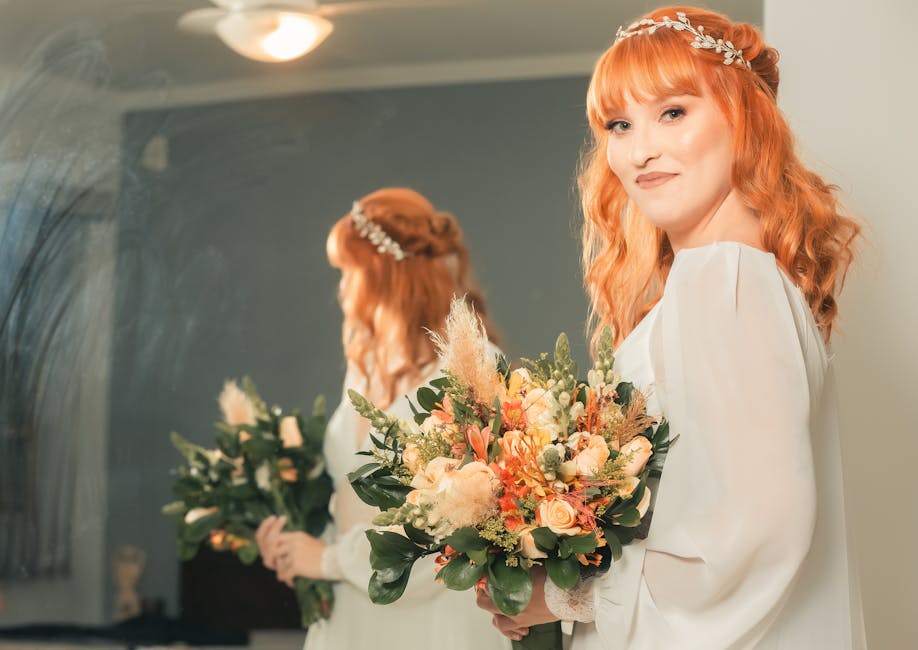 A smiling bride holding a large bouquet of fresh flowers, including white roses, eucalyptus leaves, and other delicate blooms, wrapped with a red ribbon. She is seated indoors at a wedding venue with