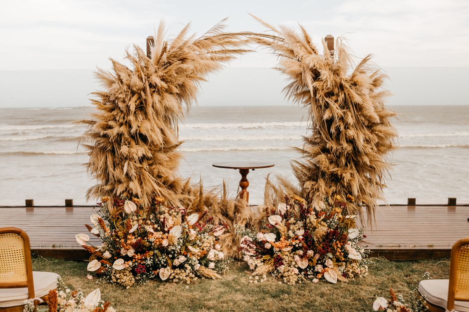 A floral installation featuring clusters of white orchids hanging beneath lush green fern foliage, supported by wooden structures. A vintage-style chandelier with brass arms and candle-shaped bulbs is