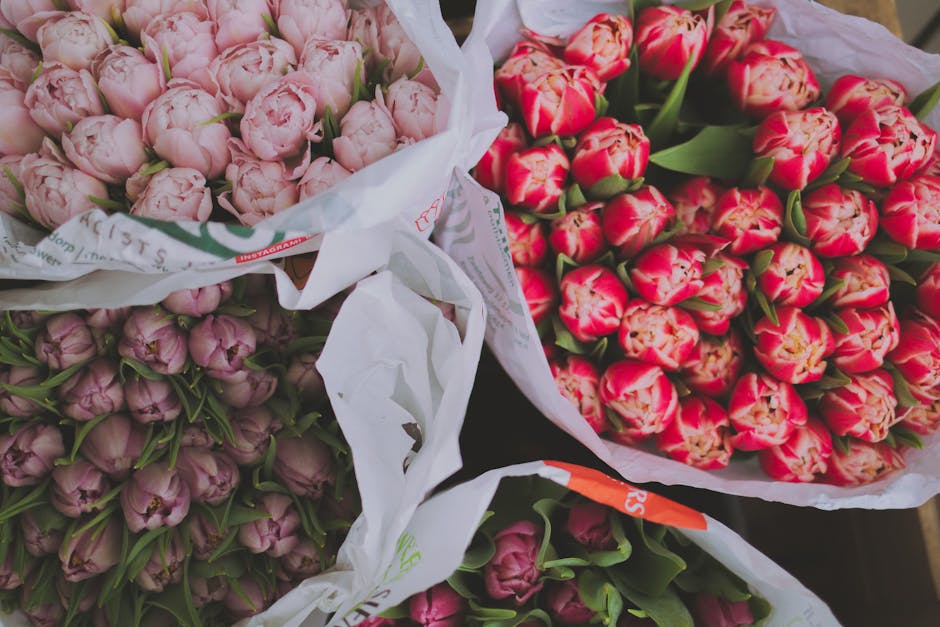 A close-up of a floral arrangement in a white, ribbed ceramic vase, featuring a mix of fresh flowers and greenery. The bouquet includes cream-colored lisianthus, a pink rose, deep purple chrysanthemum
