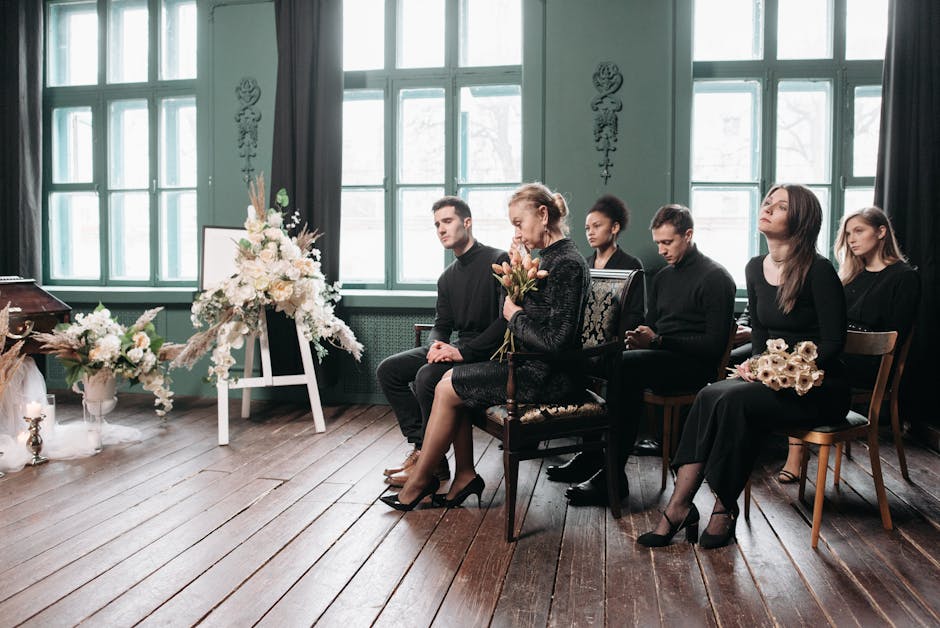 A wooden casket adorned with floral arrangements and surrounded by white candles and tall dried beige grasses, set on a surface draped with soft, sheer fabric. The floral displays include white roses,
